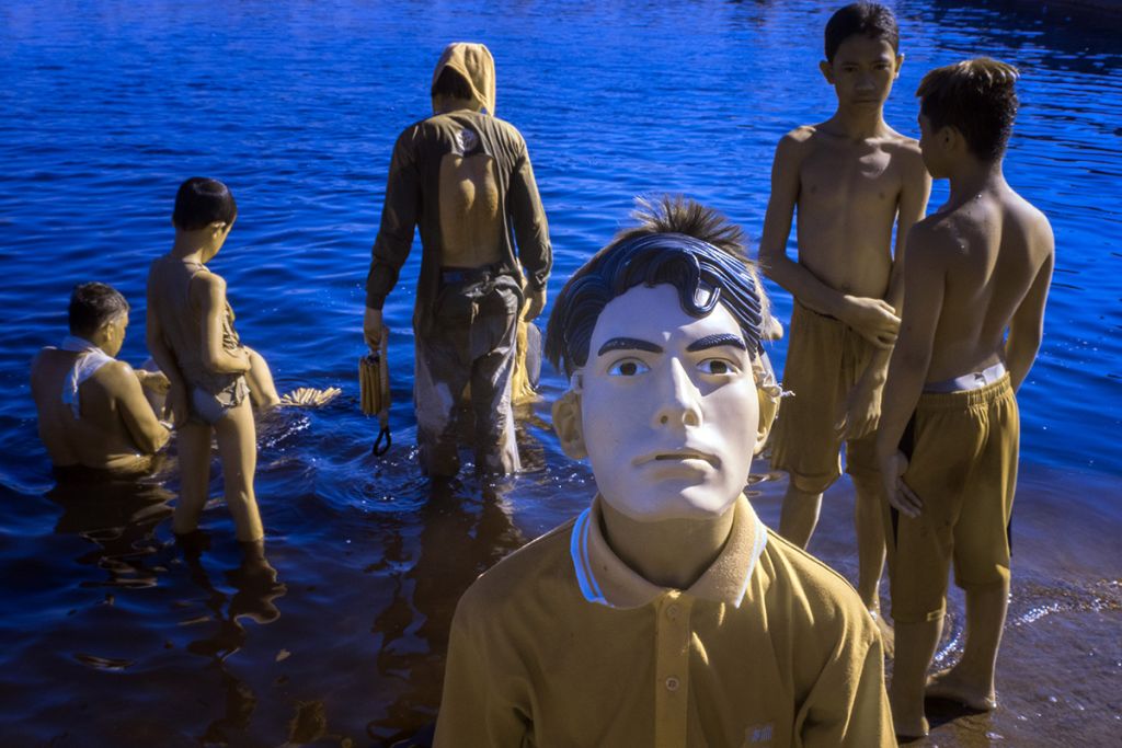 Des personnes ont les pieds dans l'eau. En face une autre personne regarde l'objectif avec un masque sur la tête.