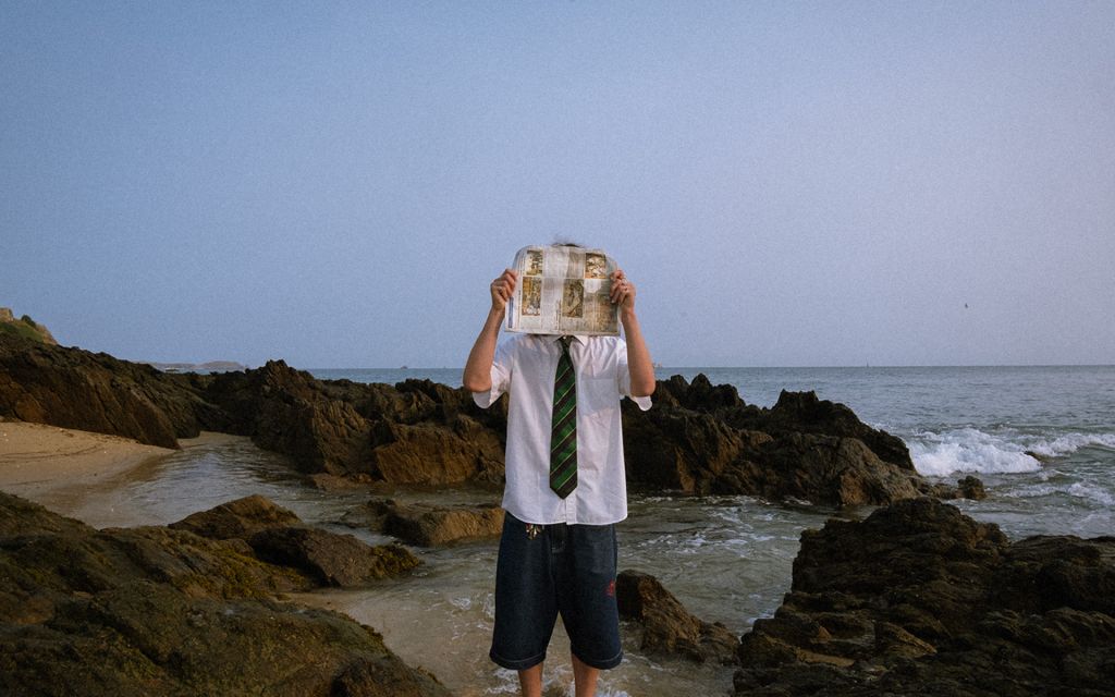 Une personne en short, chemise blanche et cravate a un journal devant le visage, près de la mer.