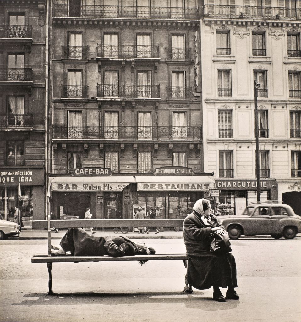 Boulevard du Montparnasse, Paris, mars 1956. Un double banc avec une dame assise au bout et un homme allongé de l'autre côté.