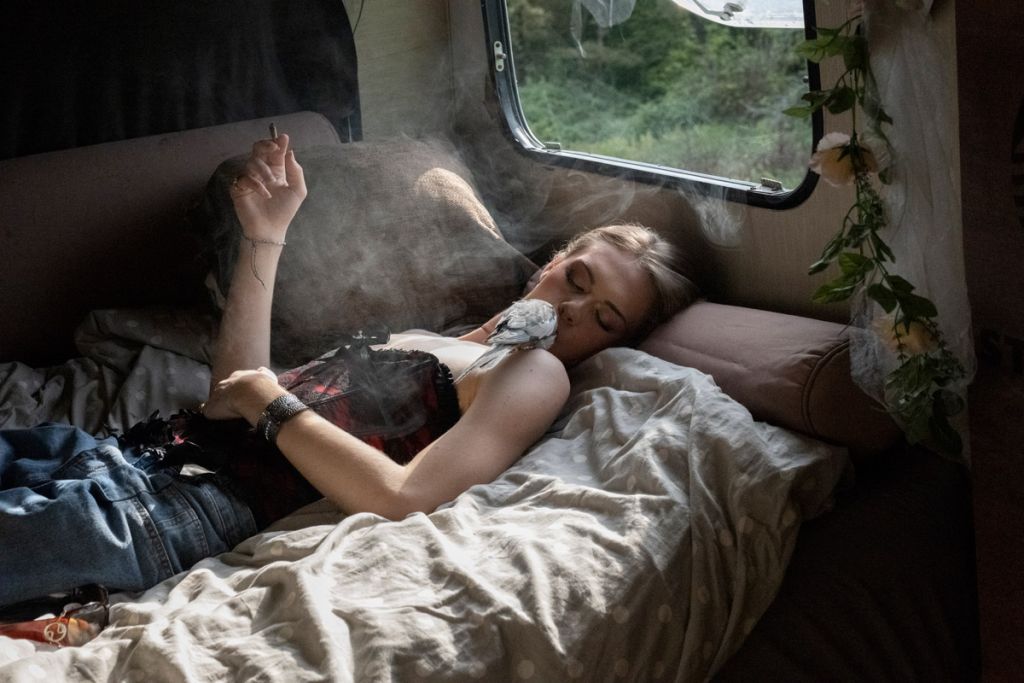 Une jeune femme est allongée sur un canapé dans un mobil-home, une cigarette à la main et un oiseau sur l'épaule