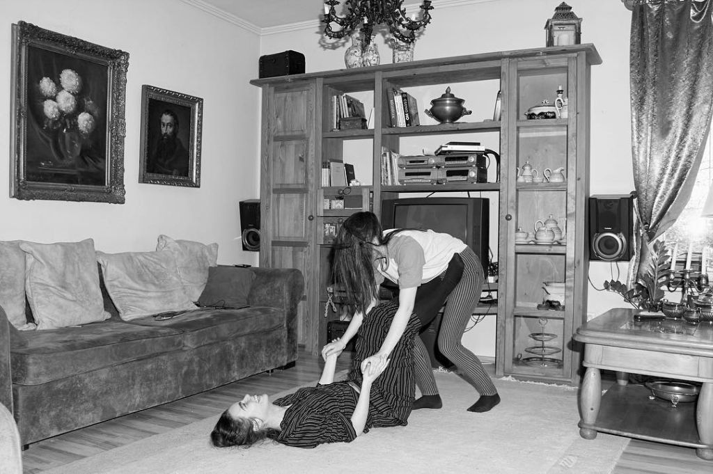 Deux jeunes femmes jouent dans un salon, l'une est allongée sur le sol les jambes tendues en l'air pour soulever le corps de la seconde.