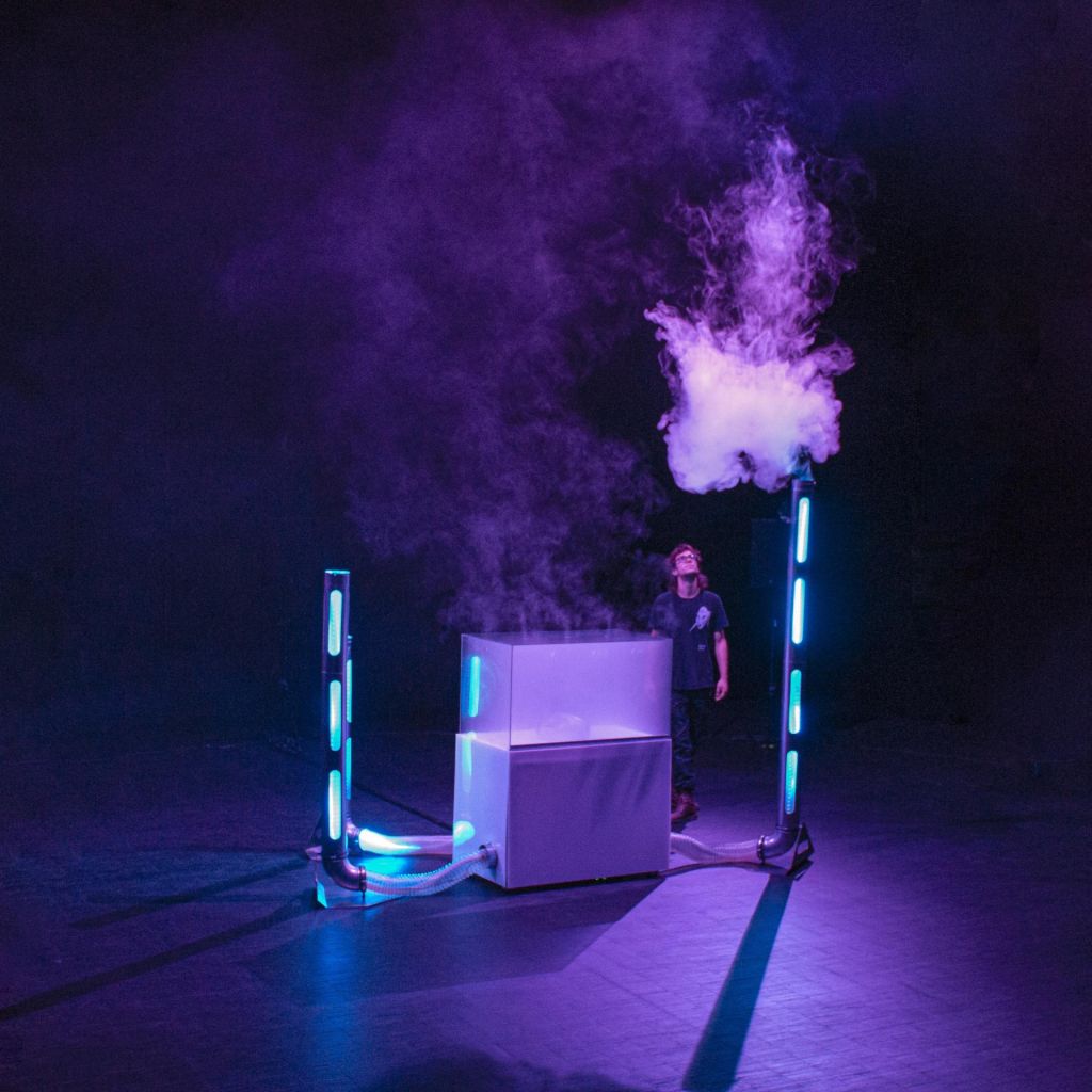 Un spectateur au pied d'une installation constituée de colonnes lumineuses et d'un aquarium observe un nuage de fumée.