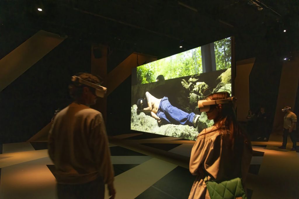 Des spectateurs, un casque VR sur la tête, dans une salle d'exposition autour d'un écran où un homme cagoulé et torse nu dort.