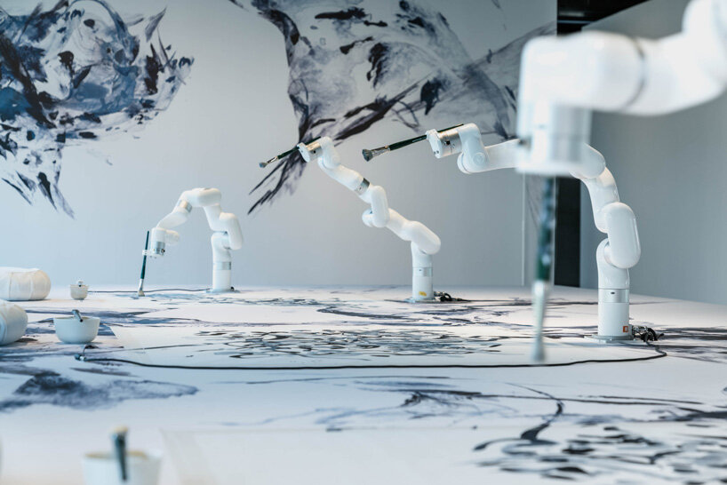 Three white robotic arms drawing on the wall and on a table.