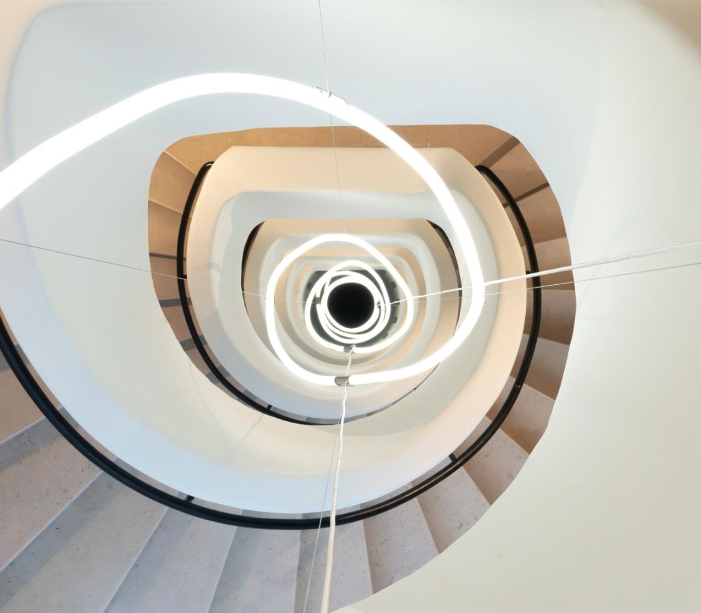 Une spirale lumineuse accompagne les mouvements d'un escalier blanc en colimaçon. 