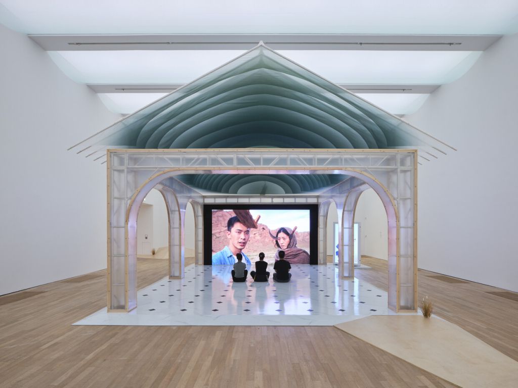 Dans une salle d'exposition, des halles ont été recréées sous lesquelles trois spectateurs regardent assis un film projeté sur grand écran.