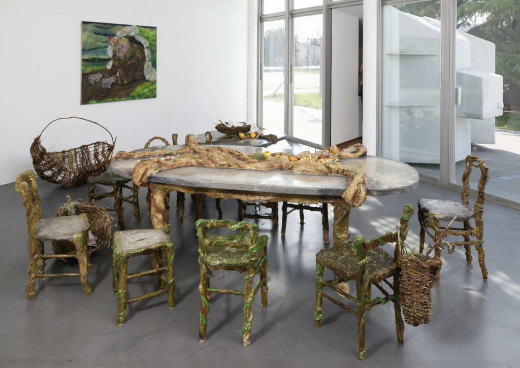 Salle à manger ouverte sur l'extérieur où les chaises sont recouvertes d'algues et où une forme monstrueuse en résine est posée sur la table.