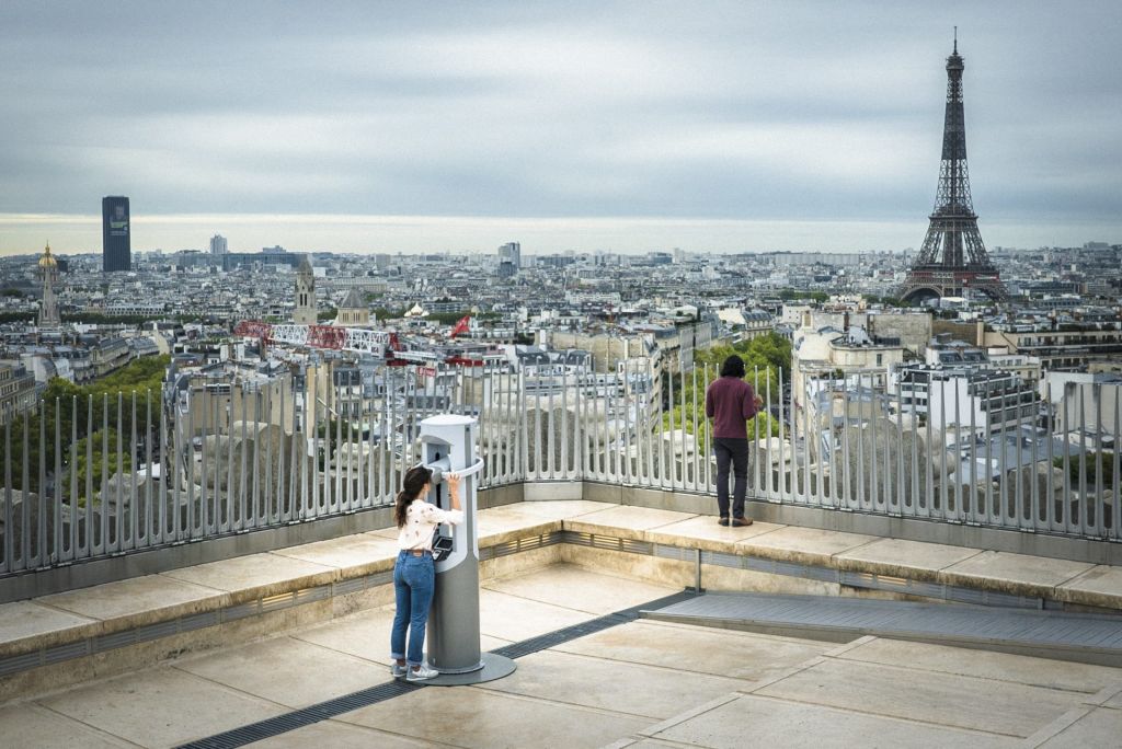 Une jeune femme depuis un toit de Paris observe l'Arc de Triomphe à l'aide d'une borne en forme d'un périscope.