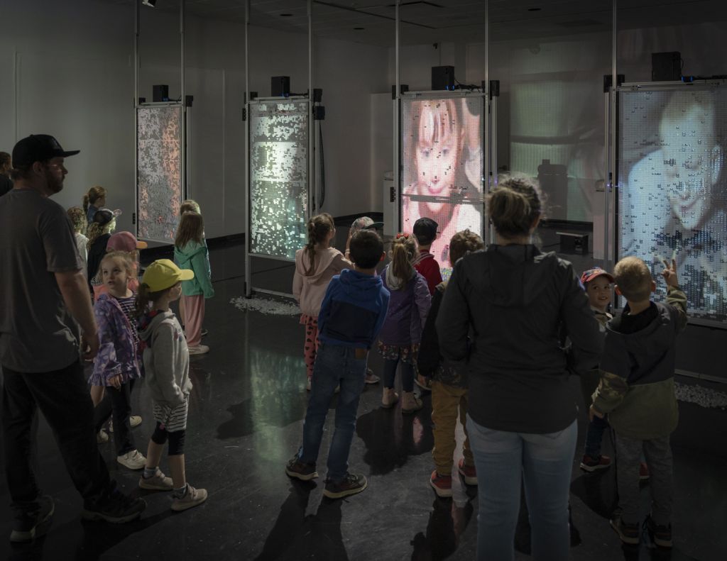 Dans une salle d'exposition, un jeune public fait face à quatre écrans sur lesquels sont projetés leurs visages pixelisés.