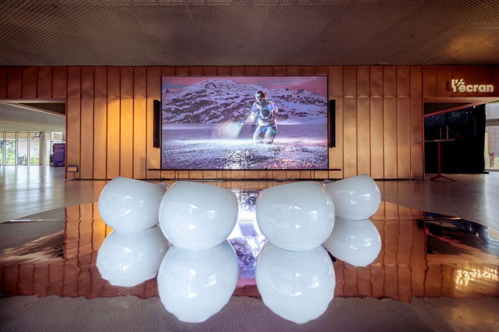 Dans une salle d'exposition, un écran géant projette un jeu vidéo avec des sièges blancs devant.