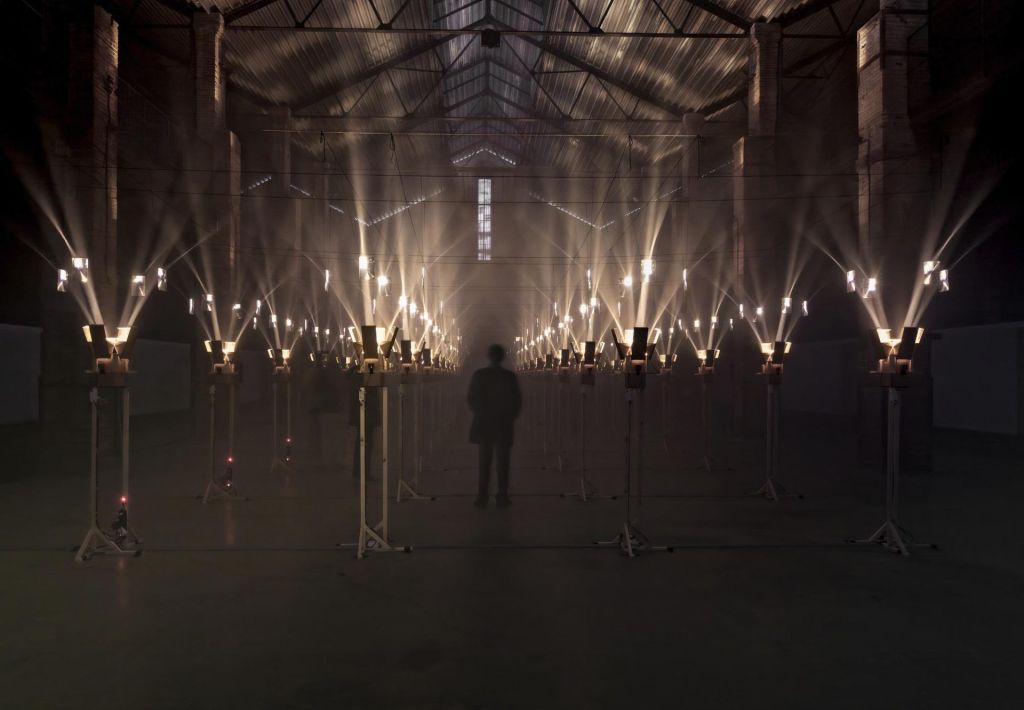 Un homme dans une église, dont l'ombre semble disparaître au milieu de dizaines de mats lumineux.