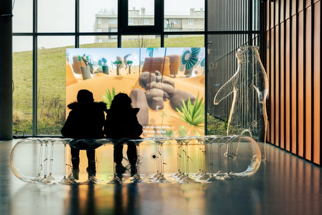 Deux enfants posés sur des siègles transparents joue à un jeu vidéo dans un lieu d'exposition.