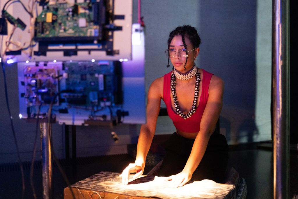 Une jeune femme métisse assise sur ses genoux en train de se servir d'un clavier en forme de tapisserie face à un écran informatique.