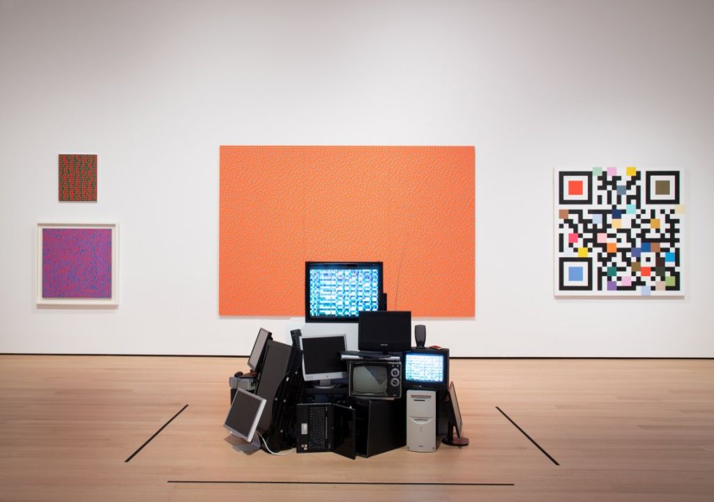 Une installation en forme d'amoncellement d'ordinateurs est positionnée au centre d'un espace musée, face à trois peintures pixelixées.