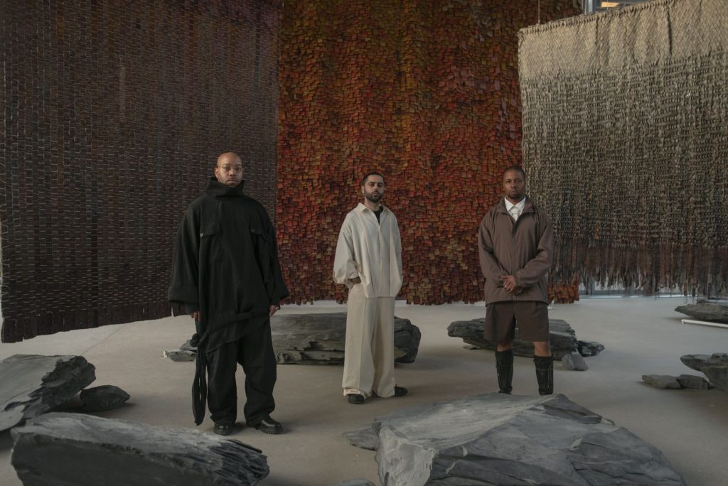 Au sein d'une installation où sont dispersés divers rochers, trois hommes se tiennent debout.