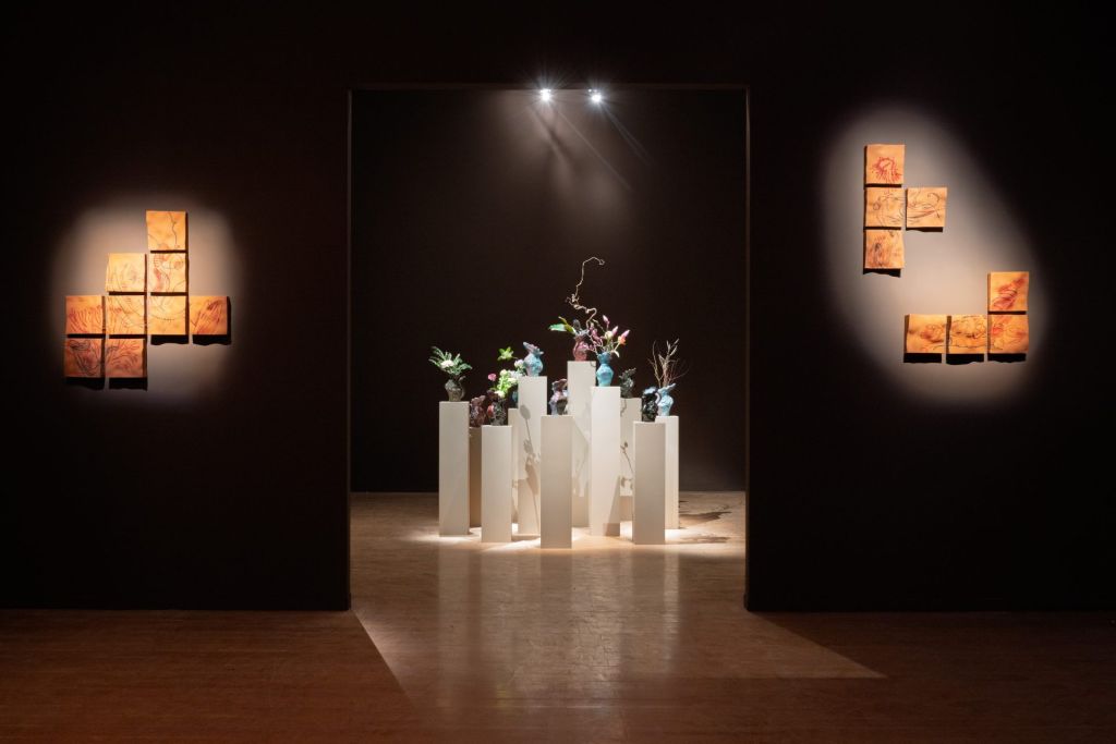 Dans un espace d'exposition, une installation faite de vases en céramique se tient au centre de la pièce, encadrée par des murs noirs sur lesquels ont été fixées des peintures rupestres.