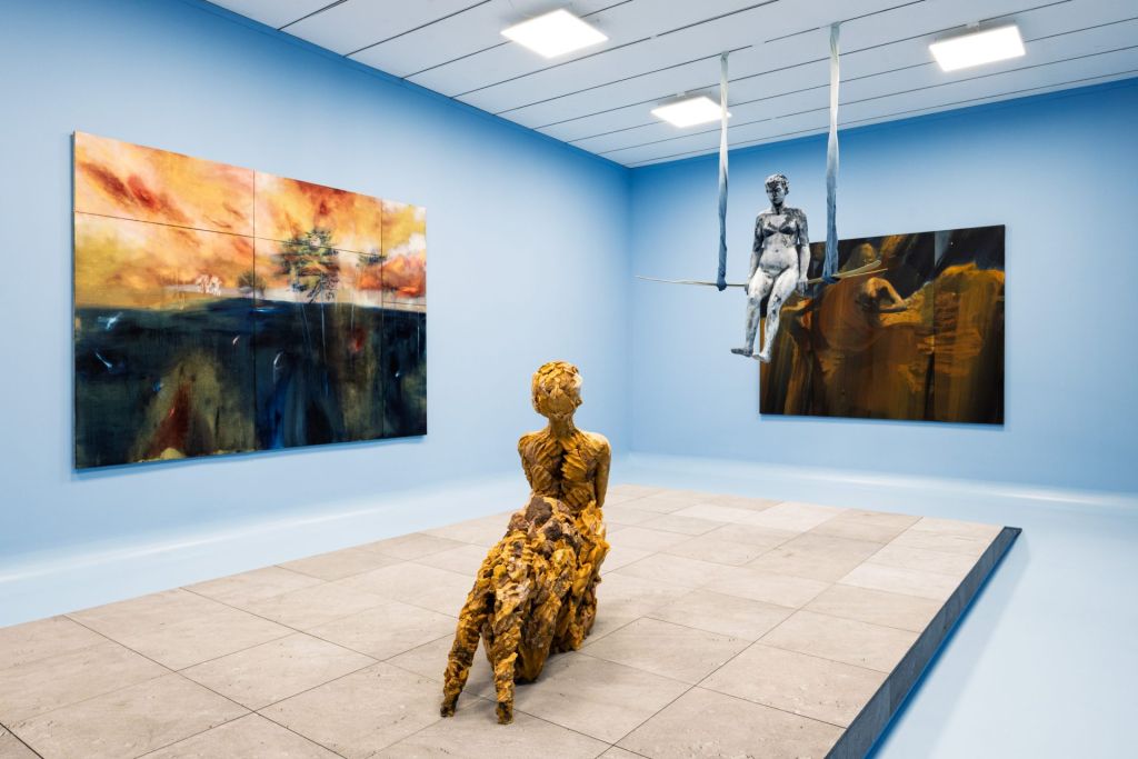 Installation dans une pièce bleue où une chimère est posée au centre de l'espace, sur du carrelage, tandis que des tableaux sont exposés tout autour.