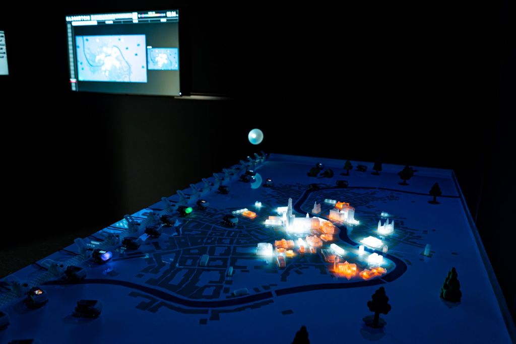 Dans le noir, une maquette reproduit des écosystèmes artificiels au sein desquels se déplaces de petits robots lumineux. 