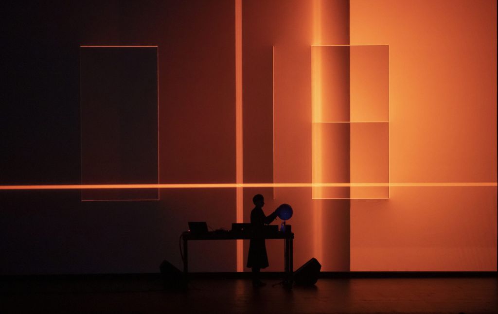 Une femme mixe sur scène avec une mappemonde devant un écran fait de formes architecturales orange.