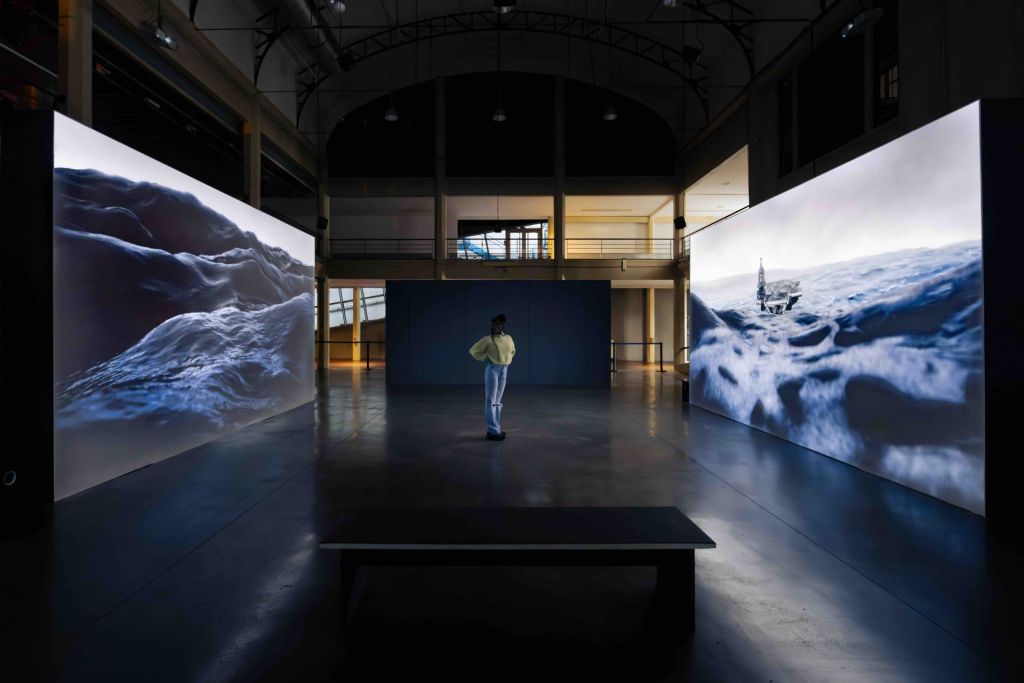 Une jeune femme se tient dans une salle d'exposition, au milieu de deux écrans géants projetants des images d'une tempête en mer.