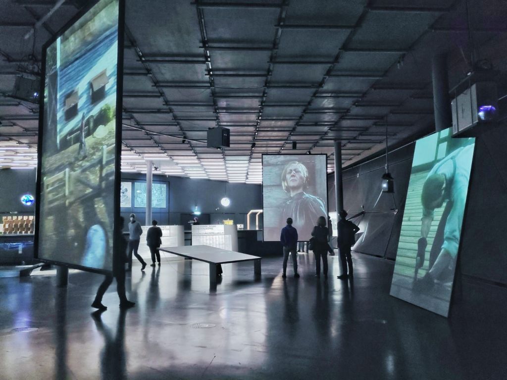Trois grands écrans vidéo diffusent un film vidéoludique dans une salle d'exposition.