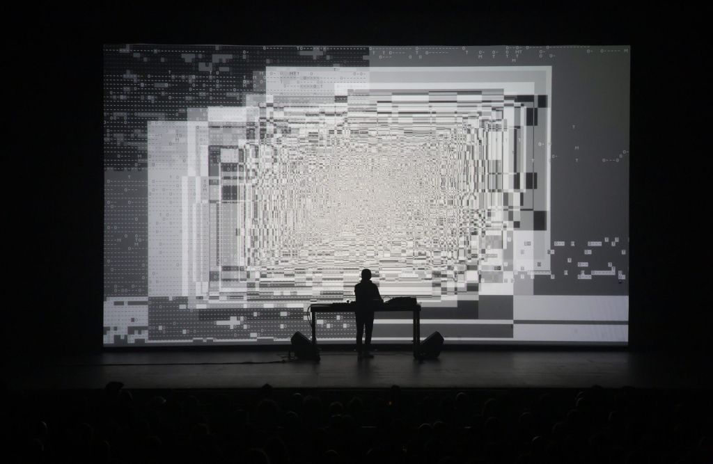 Un homme sur scène en train de mixer pendant que des images algorithmiques sont projetées sur un écran.