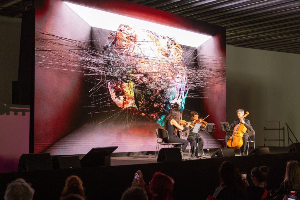 Trois musiciens à cordes performent sur scène pendant qu'un écran géant derrière eux reproduit graphiquement leurs notes.