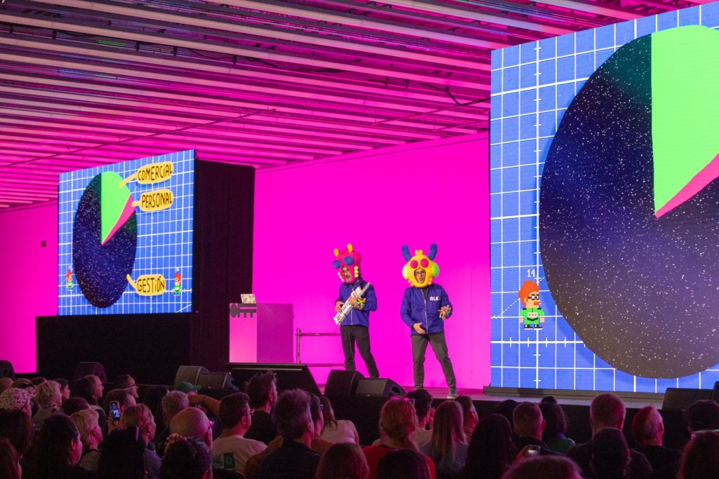 Deux hommes avec un masque de héros de mangas sur la tête performent sur scène pendant que des écrans diffusent des images à l'esthétique héritée des jeux vidéo.